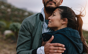 A couple stands outside, the woman leaning her head on a mans chest as he places his hand on her upper arm.
