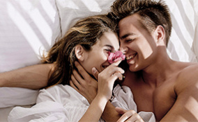 A man and woman cuddle in bed, their faces close together and a flower in the woman's hand.