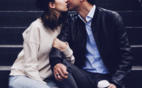 A man and woman kiss and embrace while sitting on steps outside.