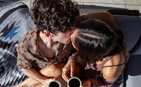 A man and and woman kiss while sitting down with cups of coffee in their hands.