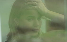 A woman looks out a green-tinted window and presses her hand to her forehead.