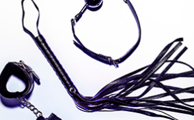 A leather whip lays between a pair of bondage cuffs and a ball gag.