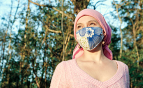 A woman in a cloth mask and head wrap stands in the sun with trees behind her.