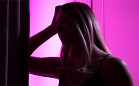 A pink light shines from behind a woman who leans her elbow against a wall and rests her head in her hands.