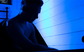 A shadowy figure sits in bed with a blue light shining behind.