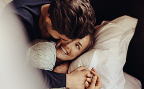 A couple cuddle in bed while smiling at each other.